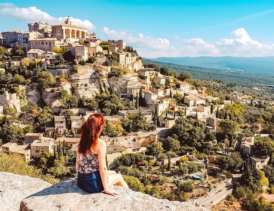 A girl with red hair perches on a rock in front of the village of Gordes France